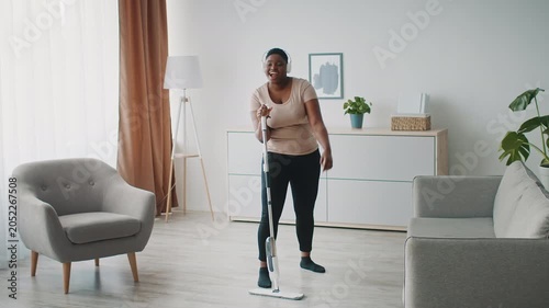 Happy Black Woman Engaging In Cleaning With Mop In Bright Modern Living Room, Smiling And Enjoying Tidying Up. Home Setting Emphasizing Cleanliness And Organization, Home Cleaning Concept.