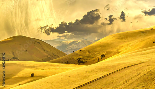 Golden rolling hills and distant mountains under a dramatic cloudy sky, suggesting a journey or exploration