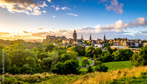 Historic Edinburgh skyline at sunset with Arthur's Seat and cityscape, dramatic clouds, golden hour light, lush green park in foreground