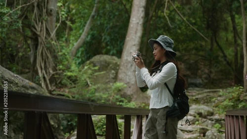 Asian woman taking pictures with a vintage camera while walking on a wooden bridge in the jungle