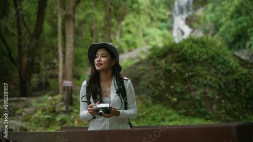 Young asian woman taking pictures with a vintage camera while hiking in a lush green forest with a waterfall