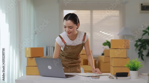Young Asian woman running a small business from home, writing down order details while working on her laptop surrounded by cardboard boxes