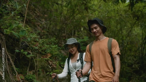 Happy young asian couple hiking together in a lush tropical forest. They are using hiking poles and carrying backpacks for their adventure