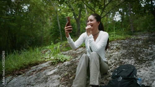 Asian woman making a video call on her smartphone while sitting on a rock in the lush green forest