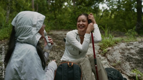 Young woman using a portable fan to cool her friend down while resting from a hike in the forest