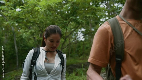 Young Asian man assisting his weary girlfriend as she rests on a large rock while trekking through a dense, vibrant forest