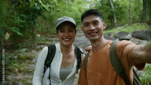 Happy asian couple taking a selfie video while hiking in a lush green forest on a summer adventure