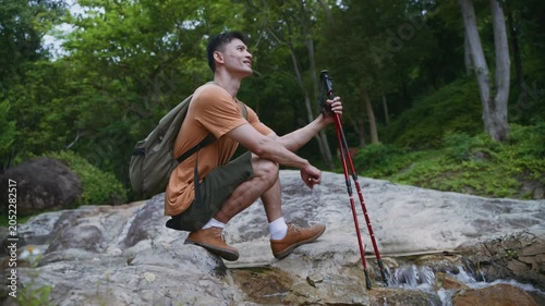 Active young asian man with a backpack and trekking poles resting by a stream in the forest, touching the cool water with his hand