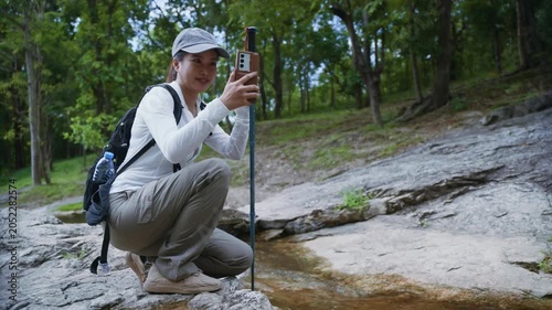 Adventurous female hiker crouching by a forest stream and taking a selfie with her smartphone while trekking in nature
