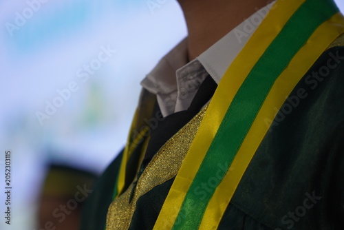 Photos Close-up of a student wearing a green graduation gown with a yellow and green sash during a commencement ceremony, representing education, achievement, and academic success
