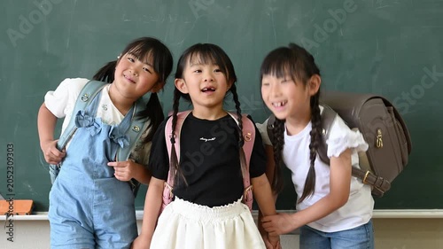 教室の黒板の前でカラフルなランドセルを背負い手をつなぐ笑顔の女子小学生たち