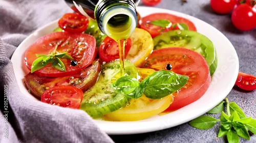 pouring olive oil from a bottle over fresh colorful heirloom tomato salad with basil on a white plate