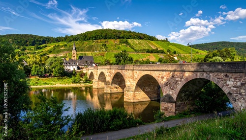 Scenic landscape featuring a stone bridge crossing a tranquil river. The scene includes a charming town and lush vineyards on a hillside