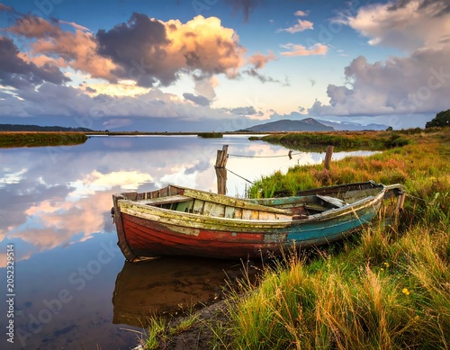 Scenic shot of an aged wooden boat resting beside a still, reflective lake mirroring a vibrant, cloudy sky at sunset