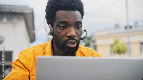 Handsome African American man with beard wearing headset talking while working on laptop outdoors. Male communicating online, sitting near modern building and enjoying remote work outside.