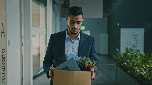 Worried Arab Man In His 30s Carrying Box Filled With Office Supplies, Personal Items, And Potted Plant, Walking Through Modern Office Space After Job Termination. Business Concept.