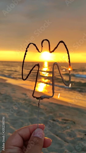 Burning sparkler in shape of angel against backdrop of sea at sunrise. Angel shaped sparkler burning on sea during sunrise sunset. Silhouette of angel in woman hand on seashore. Sparks fire. Vertical