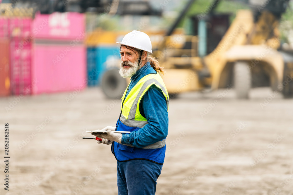 Obraz premium Construction worker in hard hat and safety vest holding a tablet at an industrial yard with containers and heavy machinery in the background.
