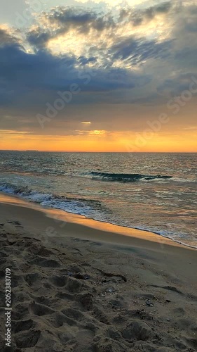 Sea waves with blue water on sand of seashore on sunset and sunrise. Yellow reflection of sunlight on surface of sea waves. Sky with dark rain clouds, horizon line and sea waves on sand. Vertical