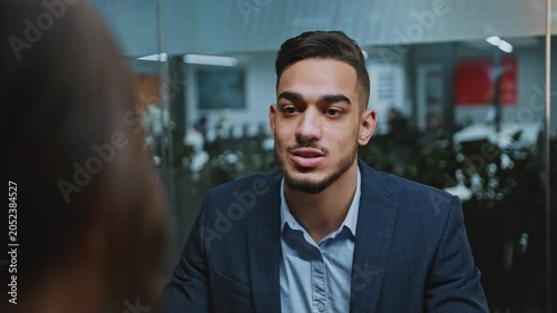 Focused Young Male Professional, In His Mid 20s, Engaging In A Serious Discussion At A Modern Office Setting. He Is Dressed In A Suit While Sitting Across From A Colleague. Business Concept.