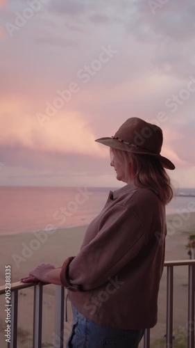 Woman in wide brim hat enjoying peaceful ocean view from balcony at pink sunset.