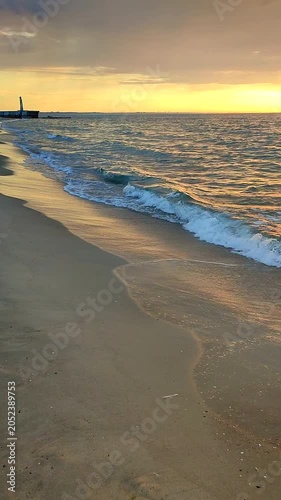 Sea waves with blue transparent water on sand of seashore on sunset and sunrise. Yellow reflection of sunlight on surface of sea waves. Sky with clouds, horizon line and sea waves on sand. Vertical