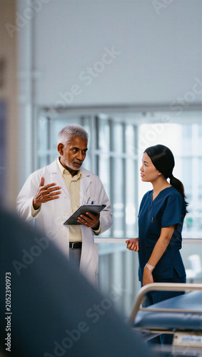 Veteran chief physician explaining digital patient charts to an attentive clinical nurse during corridor rounds.