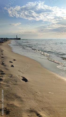 Seascape on a sunny summer day. Sea waves rolling onto the sand of a sandy beach on the seashore. Waves of a quiet calm sea, sand with footprints, blue sky with clouds and the horizon line. Vertical