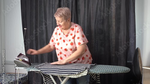 Smiling senior elderly woman ironing clothes with an iron on a board. Grandmother chores routine