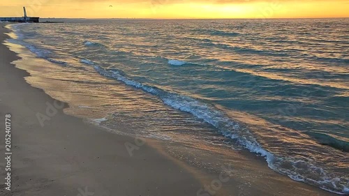 Sea waves with blue transparent water on sand of seashore on sunset and sunrise. Yellow reflection of sunlight on surface of sea waves. Sky with dark rain clouds, horizon line and sea waves on sand.