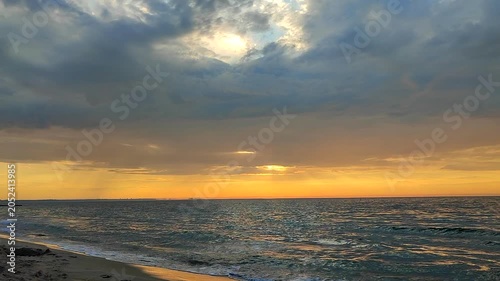 Sea waves with blue water on sand of seashore on sunset and sunrise. Yellow reflection of sunlight on surface of sea waves. Orange sky with dark rain clouds, horizon line and sea waves on sand.