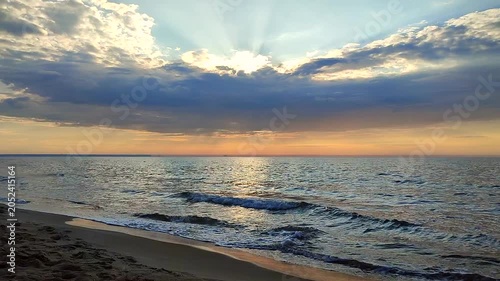 Seascape. Dark rainy clouds in blue orange sky over sea. Sunbeams from behind clouds. Sea waves on sand of seashore on sunset sunrise. Yellow reflection of sunlight on surface of sea waves.