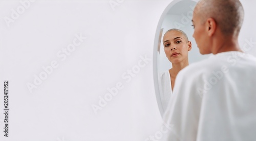 Portrait of a young woman with a buzz cut looking at her reflection in a round mirror in a minimalist white studio setting, capturing a moment of self-reflection, inner beauty, and confidence.
