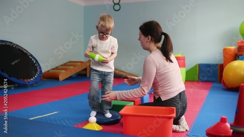 Young boy with glasses expresses excitement while sitting beside a woman on a chair in a vibrant playroom with soft flooring and play equipment