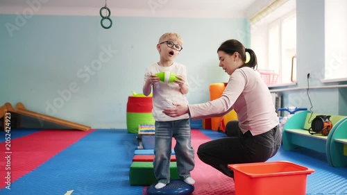 Young child wearing glasses engages with stacking toys while an adult supervises in a colorful indoor play area with soft mats and various structures