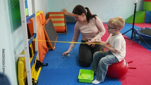 Child wearing glasses sits on a swing while a caregiver supports them in a vibrant playroom filled with various toys and equipment