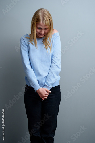Obraz na plátně A young woman wearing a light blue sweater and black pants smiles shyly while looking downwards, conveying a sense of modesty and introspection