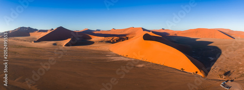 Tableau sur toile Aerial view of Dune 45 in Sossusvlei, Namibia shows red orange ridge, sharp slipface, long shadows, a road and small vehicles, early morning light across the basin