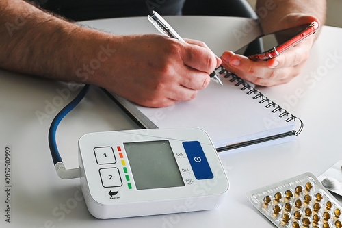 Tableau sur toile Close-up of a digital home blood pressure monitor with hands writing the reading in a notebook beside blister packs of pills illustrating hypertension self-monitoring and health tracking