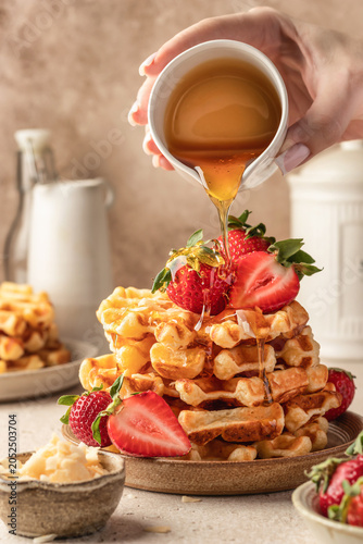 Delicious belgian waffles with strawberries on ceramic plate with honey pouring process, beige kitchen table background. Yummy sweet homemade breakfast
