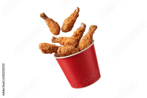 A red paper bucket filled with crispy fried chicken drumsticks, captured in mid-air with pieces floating above, isolated on a solid black background