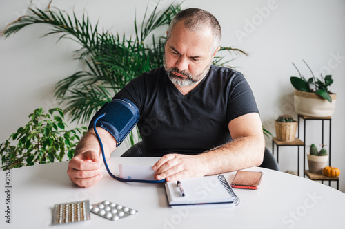 Photos Mature man measures his blood pressure with a home arm-cuff monitor at a table with blister packs of pills illustrating self care hypertension monitoring and preventive home healthcare
