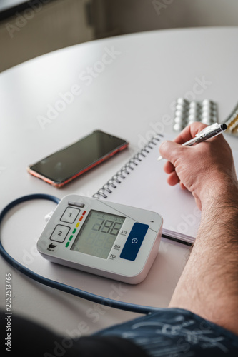 Vertical close-up of a digital home blood pressure monitor with a hand writing the reading in a notebook beside a smartphone illustrating hypertension self-monitoring at home.