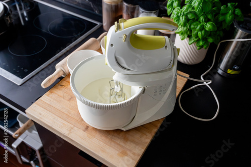Fotografie An electric hand mixer whipping fluffy white batter to soft peaks in a bowl on a wooden board in a modern marble kitchen preparing a homemade sponge cake