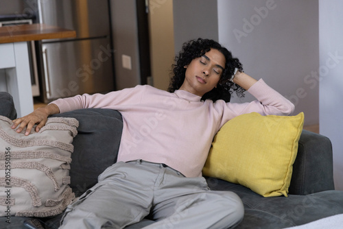 Non-binary person reclining on dark gray sofa with yellow pillow resting eyes closed near fridge