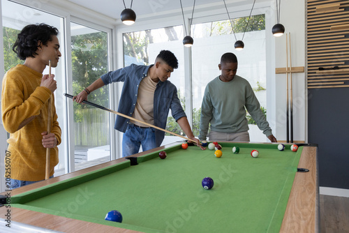 Diverse male friends playing pool on green-felt table in modern recreation room with cues