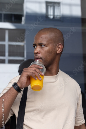 African man sipping juice from plastic dome-lid cup with straw, standing by glass facade