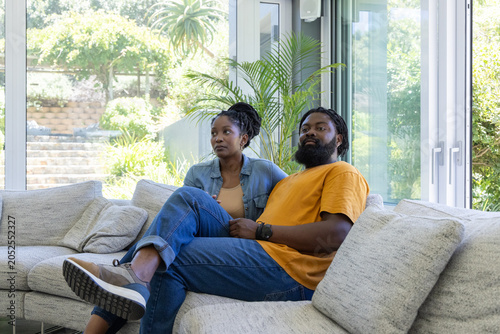 African couple sitting closely on light-gray sectional sofa near glass windows with potted palm