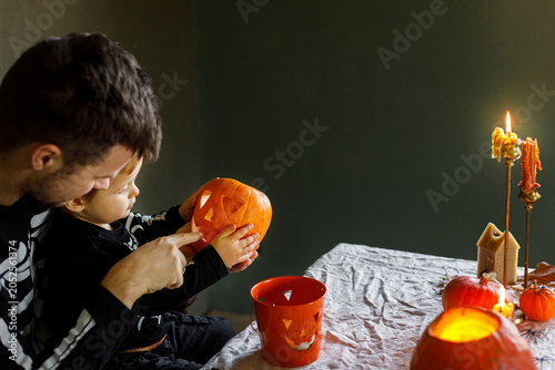 Dad and son in skeleton costumes making jack o lantern together. Happy Halloween. Father with child carving pumpkins on table with spooky decor and candles on dark background. Space for text