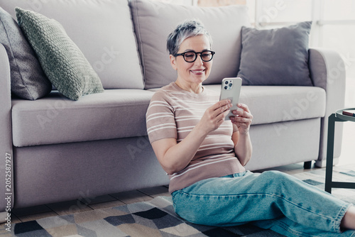 Konstfotografi Mature woman sitting on the floor by a sofa smiling and using a smartphone to te